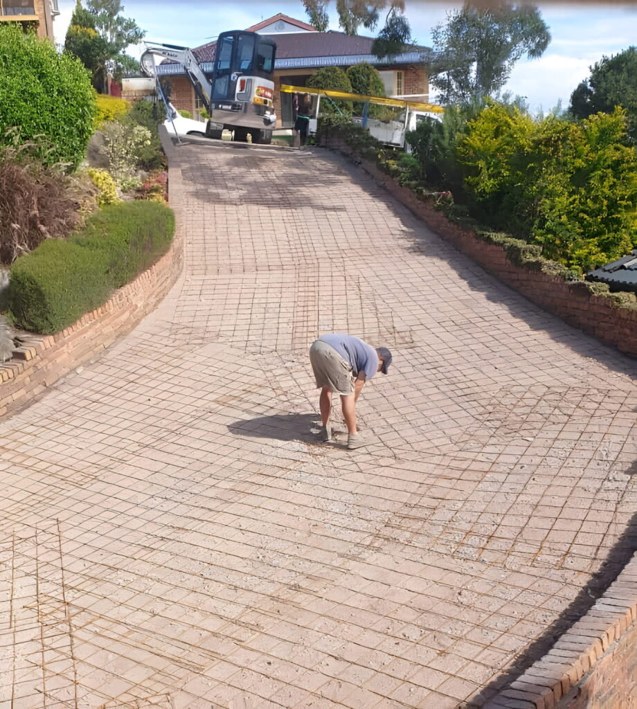 Man bending over on a grid-reinforced driveway under construction, with a small excavator parked up the slope in the background.